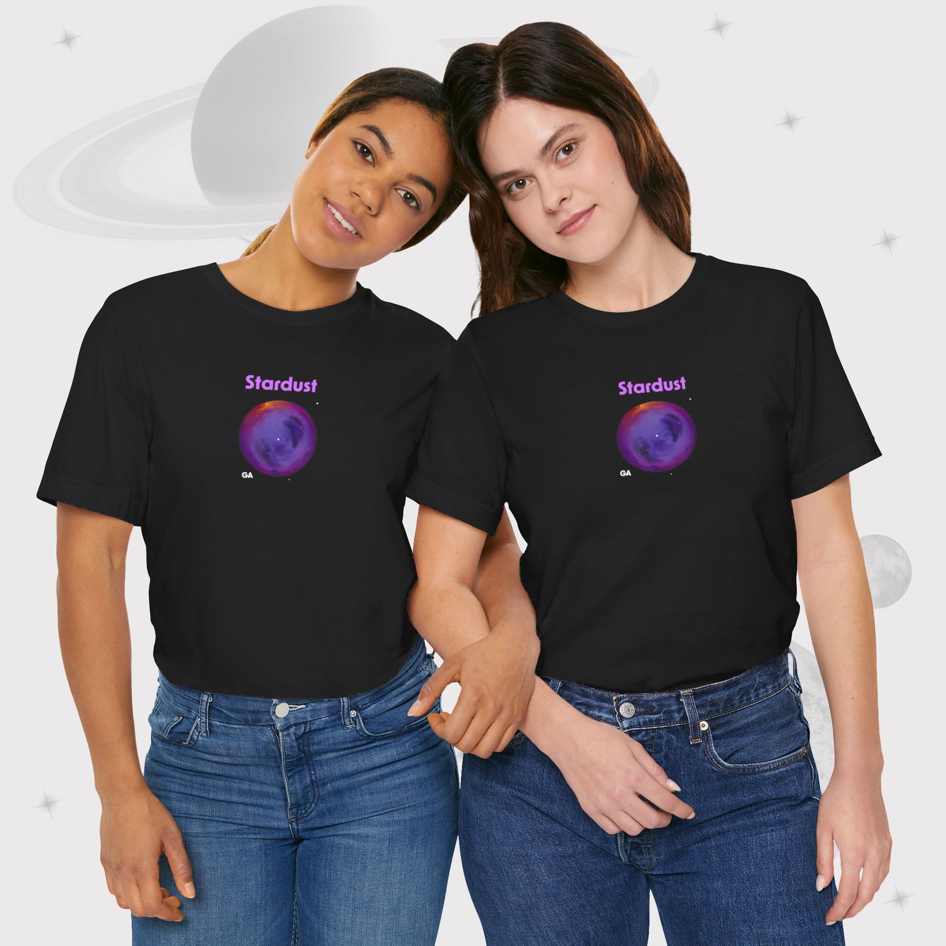 Two women wearing Going Across women's basic t-shirt in black, featuring the text ‘Stardust’ with space art of bright purple nebula by Manharleen.